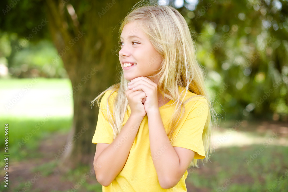 Happy beautiful Caucasian little kid girl wearing yellow T-shirt standing outdoors anticipates something awesome happen, looks happily aside, keeps hands together near face, has glad expression. Happy beautiful Caucasian little kid girl wearing yellow T-shirt standing outdoors anticipates something awesome happen, looks happily aside, keeps hands together near face, has glad expression.