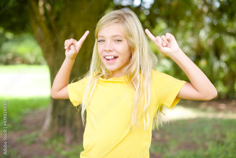 Cheerful beautiful Caucasian little kid girl wearing yellow T-shirt standing outdoors demonstrating hairdo