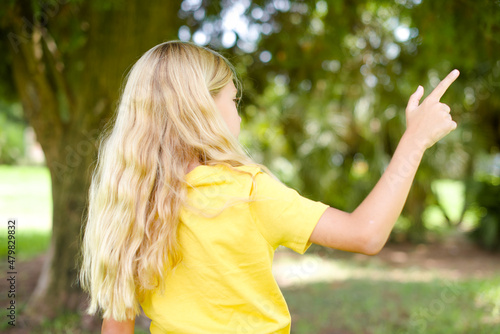 beautiful Caucasian little kid girl wearing yellow T-shirt standing outdoors pointing to object on copy space  rear view. Turn your back
