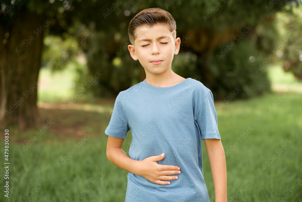 beautiful Caucasian little kid boy wearing blue T-shirt standing outdoors touches tummy, smiles gently, eating and satisfaction concept. beautiful Caucasian little kid boy wearing blue T-shirt standing outdoors touches tummy, smiles gently, eating and satisfaction concept.