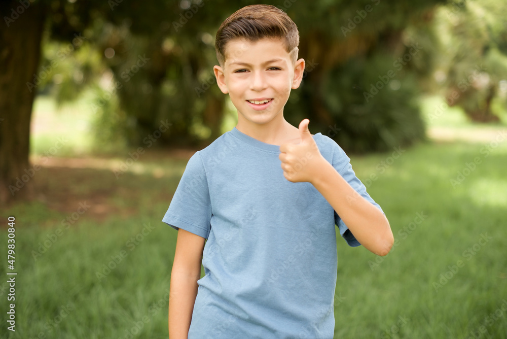 beautiful Caucasian little kid boy wearing blue T-shirt standing outdoors doing happy thumbs up gesture with hand. Approving expression looking at the camera showing success. beautiful Caucasian little kid boy wearing blue T-shirt standing outdoors doing happy thumbs up gesture with hand. Approving expression looking at the camera showing success.