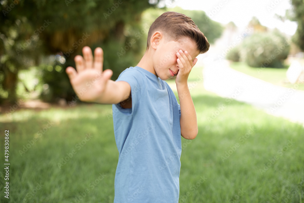 beautiful Caucasian little kid boy wearing blue T-shirt standing outdoors covers eyes with palm and doing stop gesture, tries to hide from everybody. beautiful Caucasian little kid boy wearing blue T-shirt standing outdoors covers eyes with palm and doing stop gesture, tries to hide from everybody.