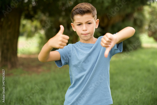 beautiful Caucasian little kid boy wearing blue T-shirt standing outdoors showing thumbs up and thumbs down, difficult choose concept