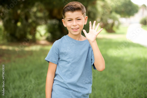 Foto beautiful Caucasian little kid boy wearing blue T-shirt standing outdoors Waiving saying hello happy and smiling, friendly welcome gesture