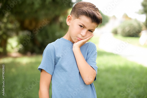 Very bored beautiful Caucasian little kid boy wearing blue T-shirt standing outdoors holding hand on cheek while support it with another crossed hand, looking tired and sick,
