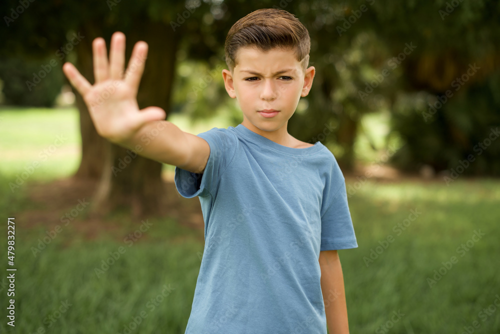 beautiful Caucasian little kid boy wearing blue T-shirt standing outdoors doing stop sing with palm of the hand. Warning expression with negative and serious gesture on the face. beautiful Caucasian little kid boy wearing blue T-shirt standing outdoors doing stop sing with palm of the hand. Warning expression with negative and serious gesture on the face.