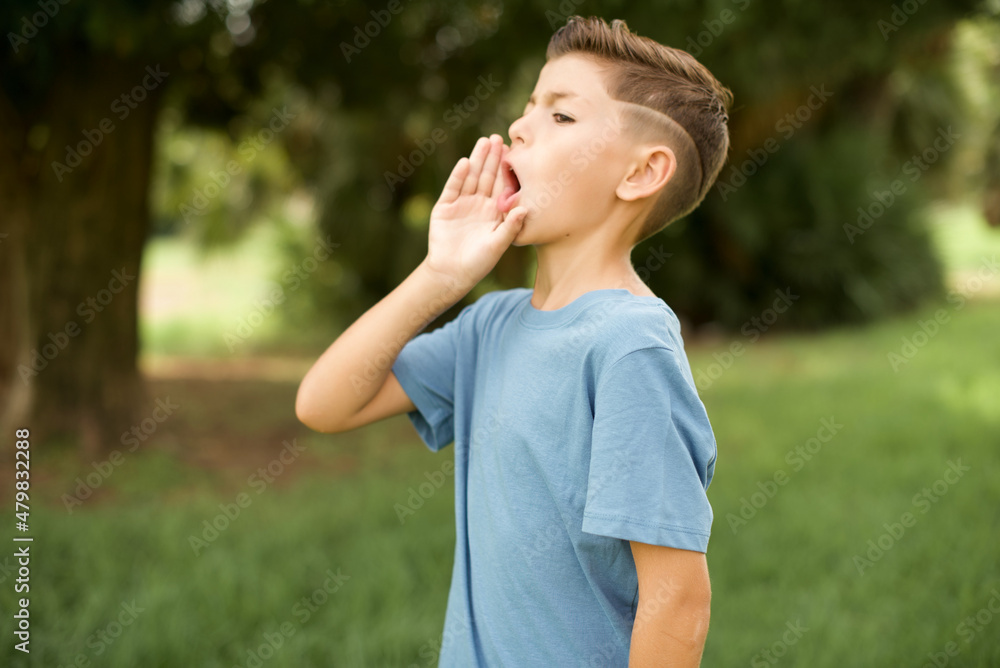 Caucasian little kid boy wearing blue T-shirt standing outdoor shouting and screaming loud to side with hand on mouth. Communication concept.