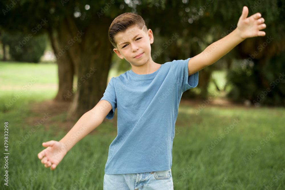 Caucasian little kid boy wearing blue T-shirt standing outdoor looking at the camera smiling with open arms for hug. Cheerful expression embracing happiness.