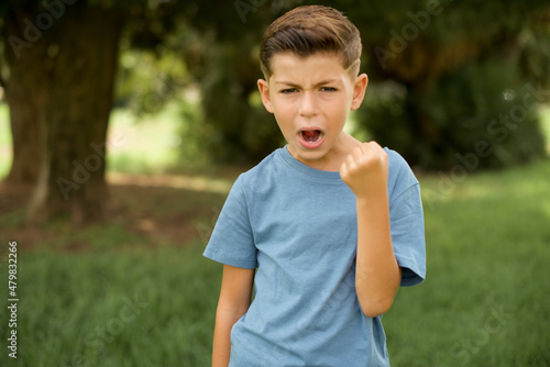 beautiful Caucasian little kid boy wearing blue T-shirt standing outdoors angry and mad raising fist frustrated and furious while shouting with anger. Rage and aggressive concept.