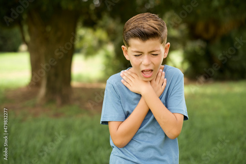 beautiful Caucasian little kid boy wearing blue T-shirt standing outdoors shouting suffocate because painful strangle. Health problem. Asphyxiate and suicide concept.
