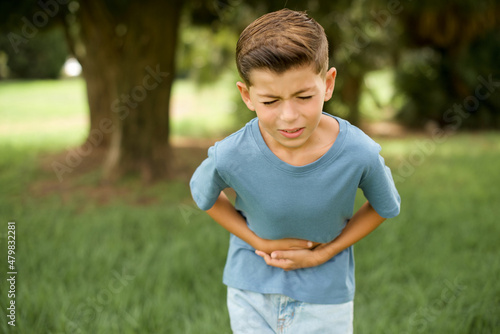 Caucasian little kid boy wearing blue T-shirt standing outdoor with hand on stomach because nausea, painful disease feeling unwell. Ache concept.