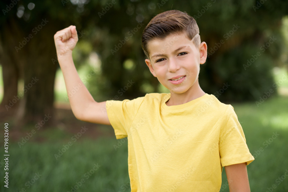 Portrait of powerful cheerful Caucasian little kid boy wearing yellow T-shirt standing outdoor showing muscles. Portrait of powerful cheerful Caucasian little kid boy wearing yellow T-shirt standing outdoor showing muscles.