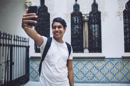 Toothy male tourist using front camera on mobile device for clicking funny photo content during getaway travelling, cheerful hipster guy shooting web vlog about international trip on vacations photo