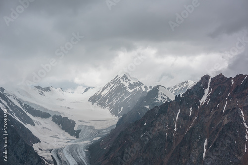 Atmospheric landscape with sharp rocks and high snowy mountain top in rainy low clouds at overcast. Dramatic gloomy scenery with large snow mountains and glacier in gray cloudy sky at rainy weather.