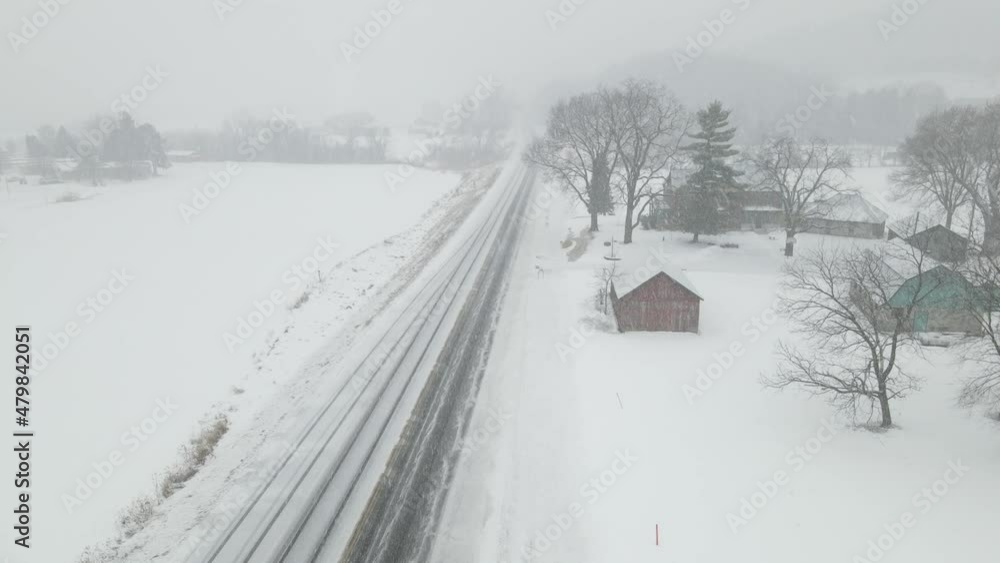 Heavy snow falling over a rural highway and farm. Snow covered road. Diminished visibility in the distance. One semi trailer traveling down the highway. Heavy snow falling over a rural highway and farm. Snow covered road. Diminished visibility in the distance. One semi trailer traveling down the highway.