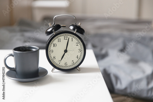 Image alarm clock with a black cup of coffee on a white bedside table in front of the bed with gray linens. The room is in beige tones.