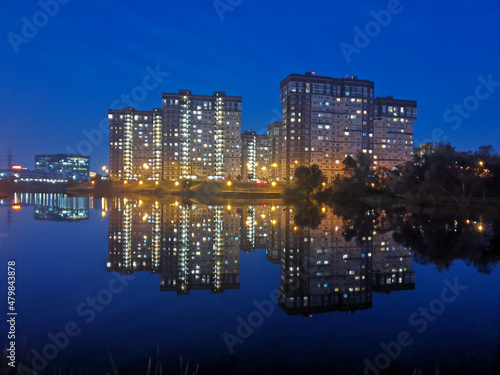 Multi-storey residential buildings with glowing windows are reflected in blue water against the background of the night sky