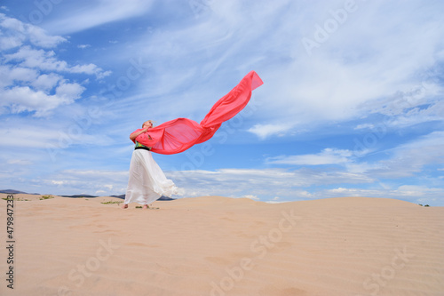 Young woman standing in sand dunes with a wide and large coral pink color textile silk scarf around her on a windy day.