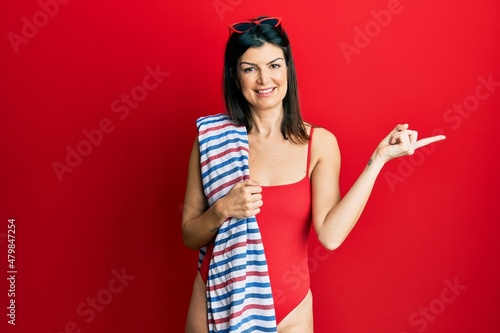 Young hispanic woman wearing swimsuit holding beach towel smiling happy pointing with hand and finger to the side