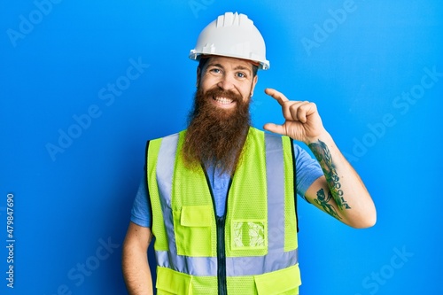 Redhead man with long beard wearing safety helmet and reflective jacket smiling and confident gesturing with hand doing small size sign with fingers looking and the camera. measure concept.