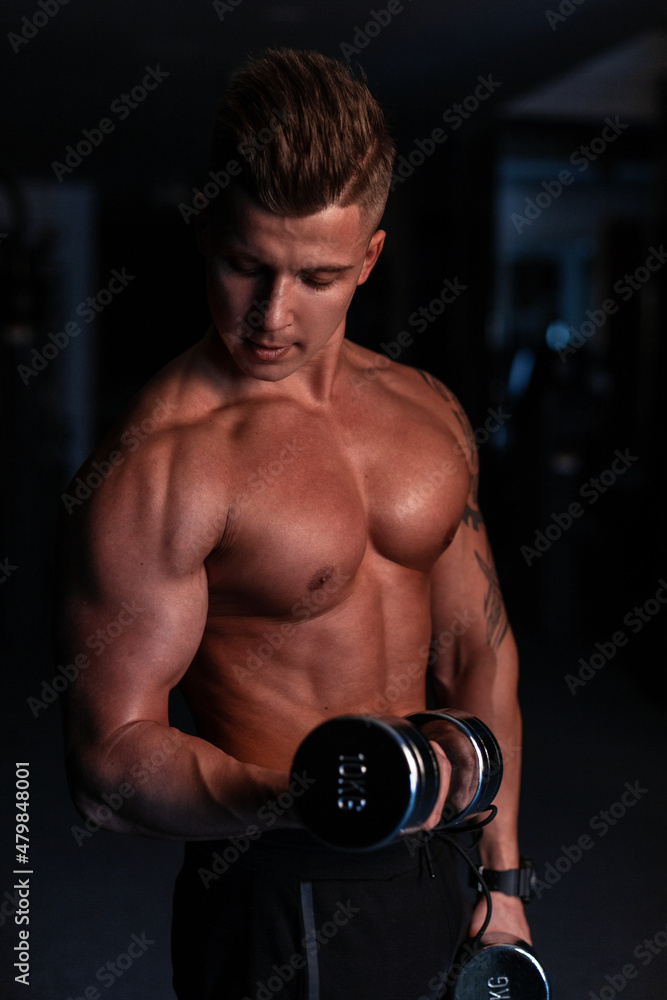 Handsome sports sexy young man with a naked  torso and muscles does an exercise with dumbbells and rocks his biceps in the gym