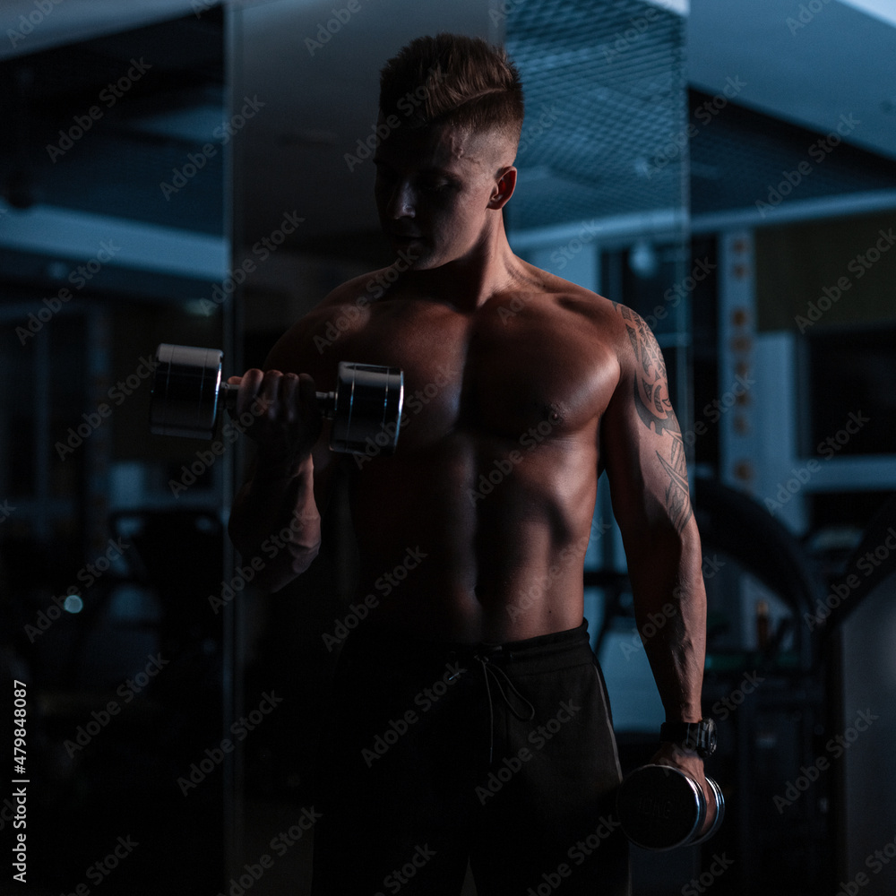 Handsome young fitness trainer man with naked torso with muscles doing workout with dumbbells in the dark in the gym