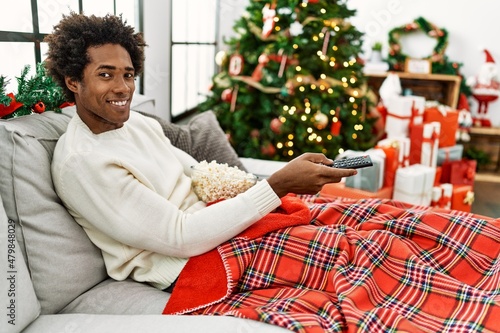 Εκτύπωση καμβά Young african american man watching movie sitting on the sofa by christmas tree at home