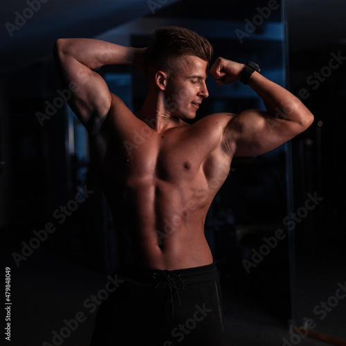 Handsome young sports man with a sexy naked torso and muscles in the gym on a dark background