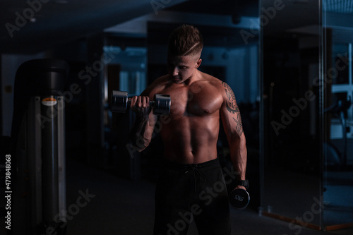 Fitness man model with hairstyle and tattoo workout with dumbbell in gym in the dark. Sports lifestyle