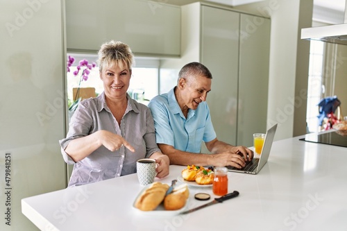 Middle age caucasian couple eating breakfast at home using laptop smiling happy pointing with hand and finger