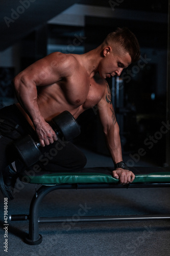 Strong athletic young man with naked torso and muscles doing dumbbell workout in the gym at night