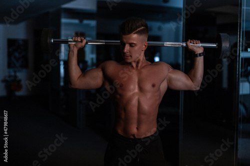 Strong handsome guy with a naked muscular body holding the bar and working out in the gym on a dark background