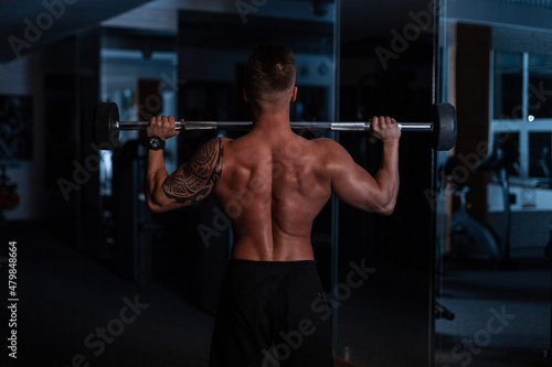 Sports man trainer with muscular back and tattoo on his shoulder doing exercises with dumbbells in the gym on a blue dark background