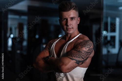 Cool young sexy man with a hairstyle and muscular body with a tattoo in a white t-shirt posing in the gym on a dark blue background © alones