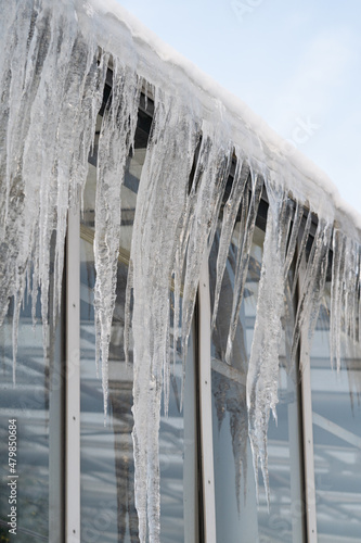 Icicles hanging on glass roof and wall in winter. Rooftop covered with ice and snow during spring thaw  melting water running down and froze at night. Roof insulation maintenance problem concept