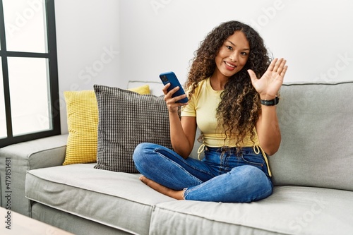 Beautiful hispanic woman sitting on the sofa at home using smartphone waiving saying hello happy and smiling, friendly welcome gesture