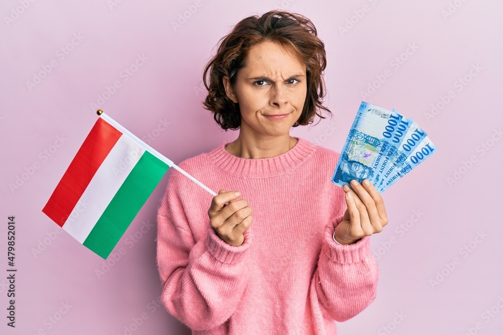 Young brunette woman holding hungary flag and forints banknotes skeptic and nervous, frowning upset because of problem. negative person. Young brunette woman holding hungary flag and forints banknotes skeptic and nervous, frowning upset because of problem. negative person.