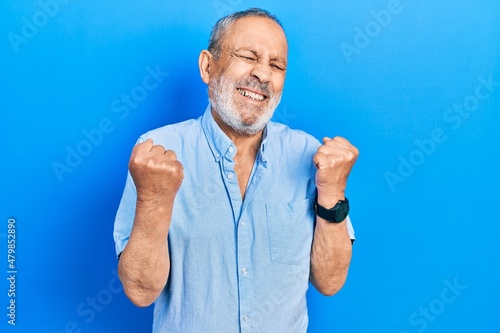 Handsome senior man with beard wearing casual blue shirt very happy and excited doing winner gesture with arms raised, smiling and screaming for success. celebration concept.