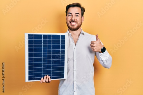 Handsome caucasian man with beard holding photovoltaic solar panel smiling happy and positive, thumb up doing excellent and approval sign photo