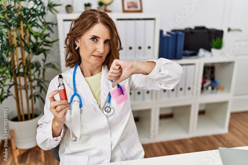 Middle age doctor woman holding electronic cigarette at medical clinic with angry face  negative sign showing dislike with thumbs down  rejection concept