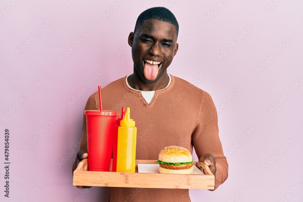 Young african american man eating a tasty classic burger and soda sticking tongue out happy with funny expression. Young african american man eating a tasty classic burger and soda sticking tongue out happy with funny expression.