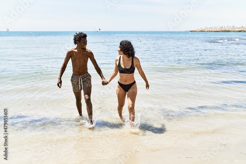Young interracial tourist couple wearing swimwear walking with hands together at the beach.