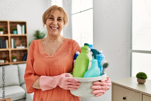 Middle age blonde woman holding cleaning products cleaning at home looking positive and happy standing and smiling with a confident smile showing teeth