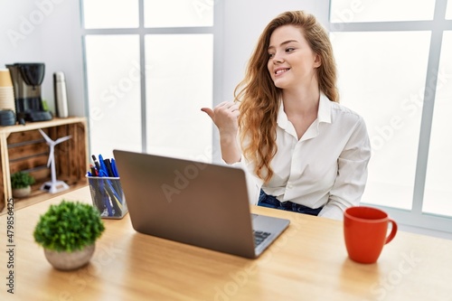 Young caucasian woman working at the office using computer laptop smiling with happy face looking and pointing to the side with thumb up.