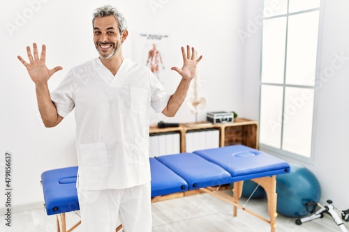 Middle age hispanic therapist man working at pain recovery clinic showing and pointing up with fingers number ten while smiling confident and happy.