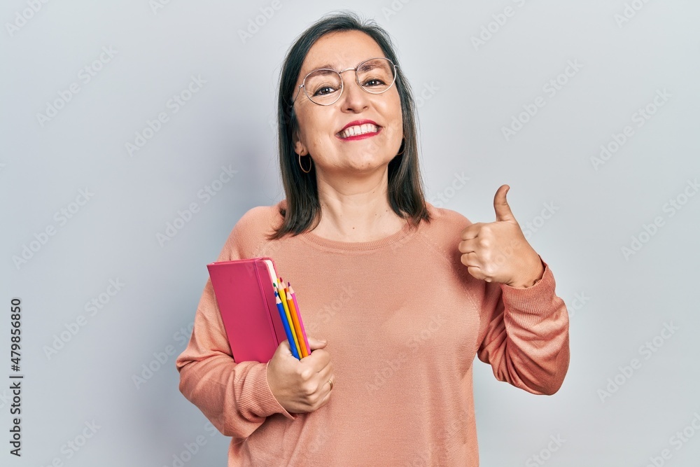 Middle age hispanic woman holding book and color pencils smiling happy and positive, thumb up doing excellent and approval sign Middle age hispanic woman holding book and color pencils smiling happy and positive, thumb up doing excellent and approval sign