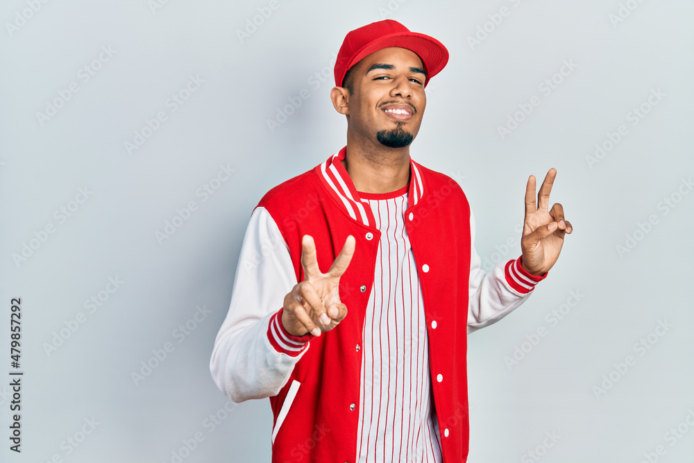 Young african american man wearing baseball uniform smiling looking to the camera showing fingers doing victory sign. number two. Young african american man wearing baseball uniform smiling looking to the camera showing fingers doing victory sign. number two.