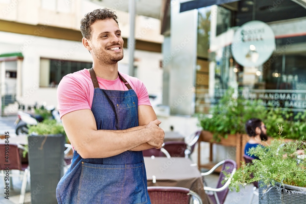 Obraz premium Young hispanic waiter wearing apron standing with arms crossed gesture at coffee shop.