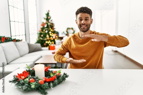Arab young man sitting on the table by christmas tree gesturing with hands showing big and large size sign  measure symbol. smiling looking at the camera. measuring concept.