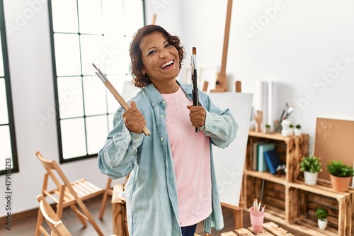 Middle age hispanic artist woman smiling happy holding paintbrushes at art studio.